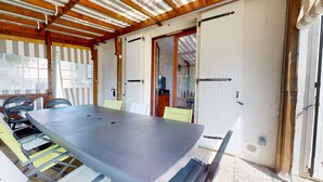A covered patio with a table and chairs, wooden beams, and a sliding glass door leading to an interior space. - Dolce Vita close to the beach - Terrace & Garden (Saint-Hilaire-de-Riez)