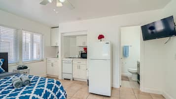 A bedroom with a bed, a ceiling fan, a TV, a kitchen area with cabinets, a refrigerator, and a bathroom with a toilet.