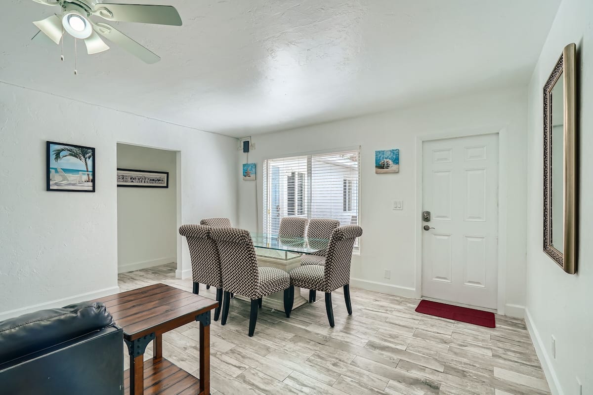 A dining area with a glass table and patterned chairs, a ceiling fan, and a door with a red doormat.