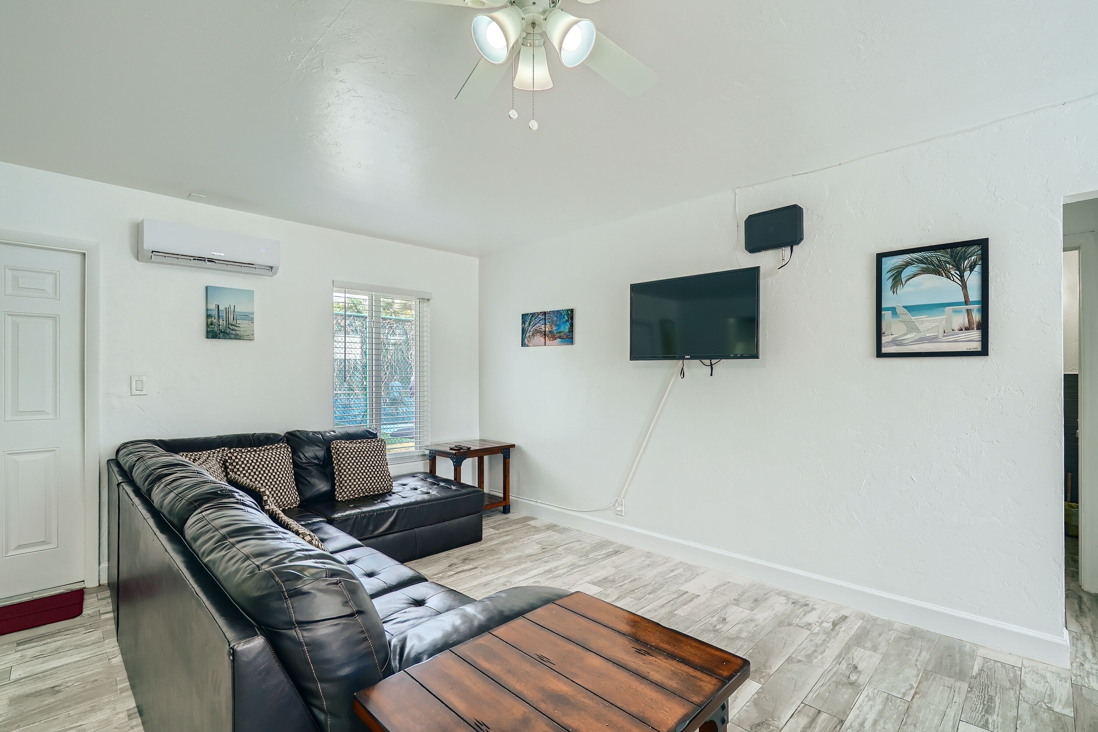 A living room with a black leather sofa, a wooden coffee table, and a ceiling fan. There is a TV mounted on the wall and a window with blinds.