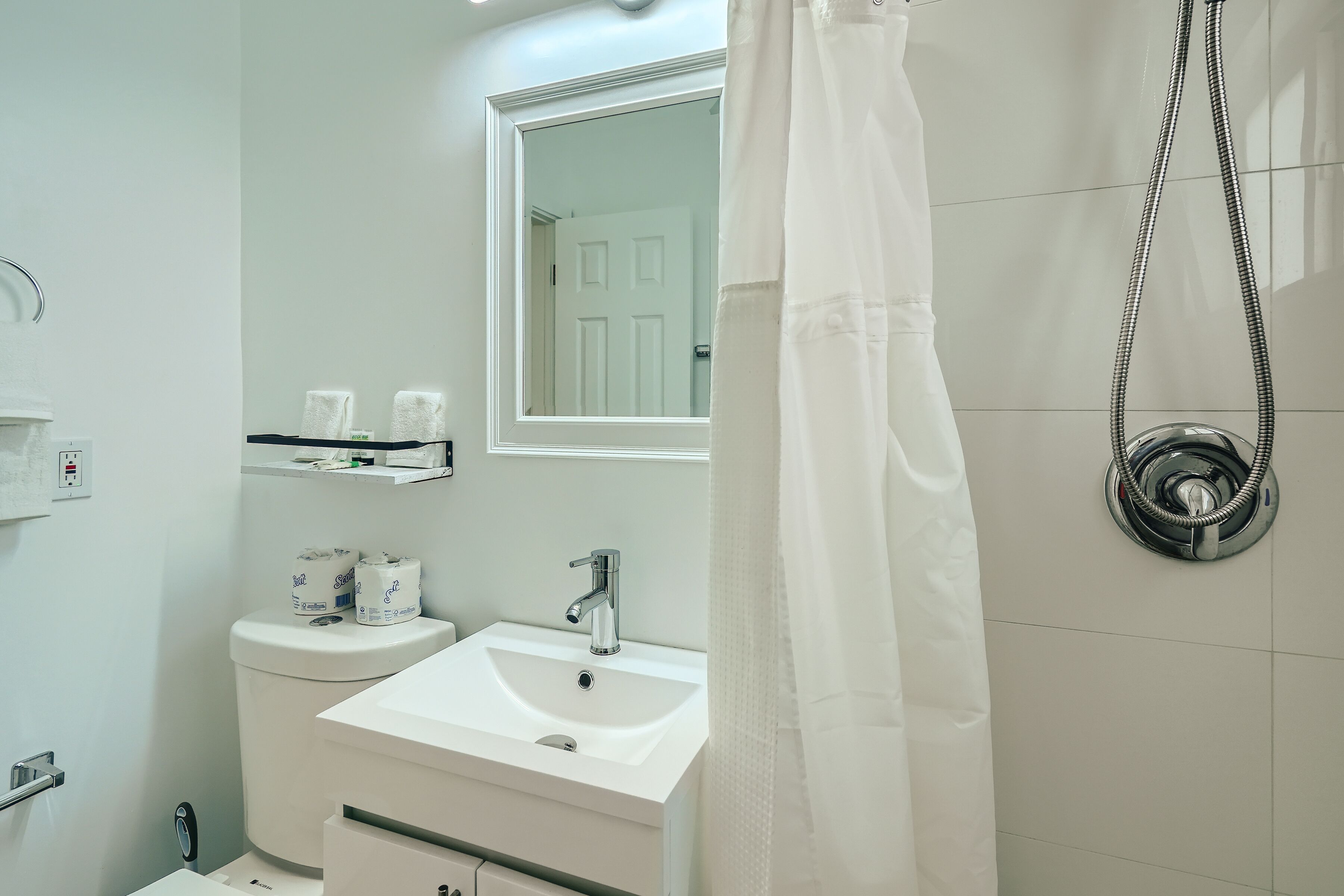 Une salle de bain avec un lavabo blanc, un miroir et des toilettes. Un pommeau de douche est fixé au mur et un rideau est partiellement tiré.