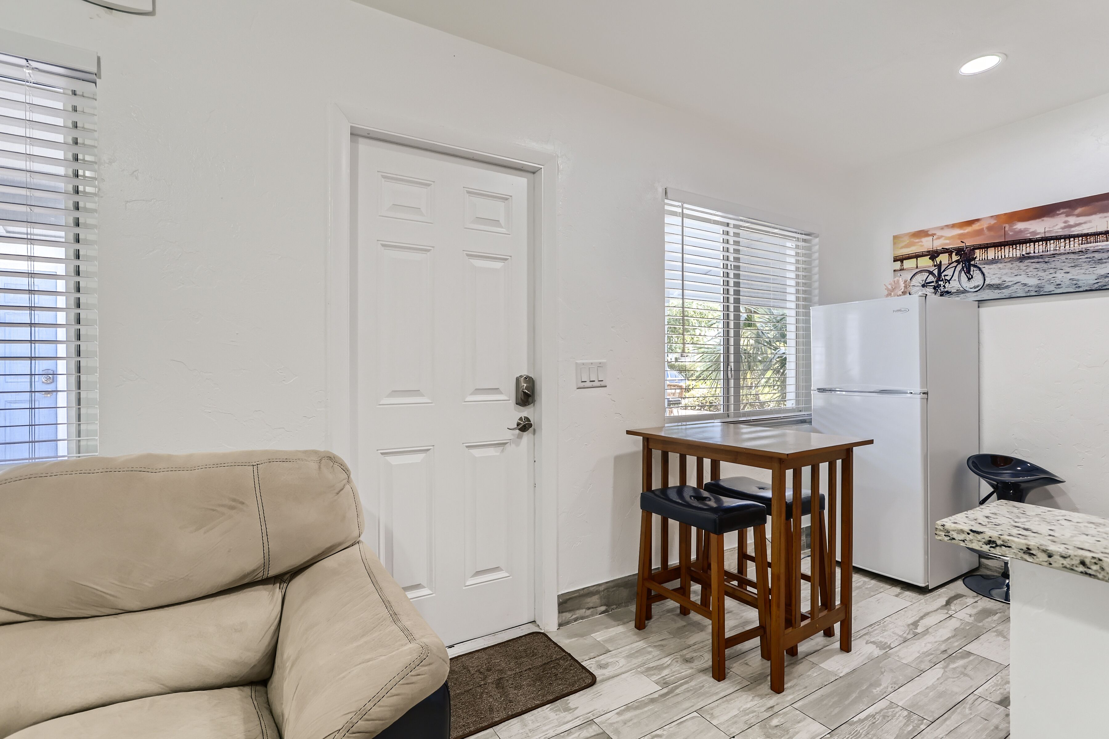 Un espace de vie compact comprenant un canapé beige, une petite table en bois avec deux chaises, un réfrigérateur et une fenêtre avec des stores.