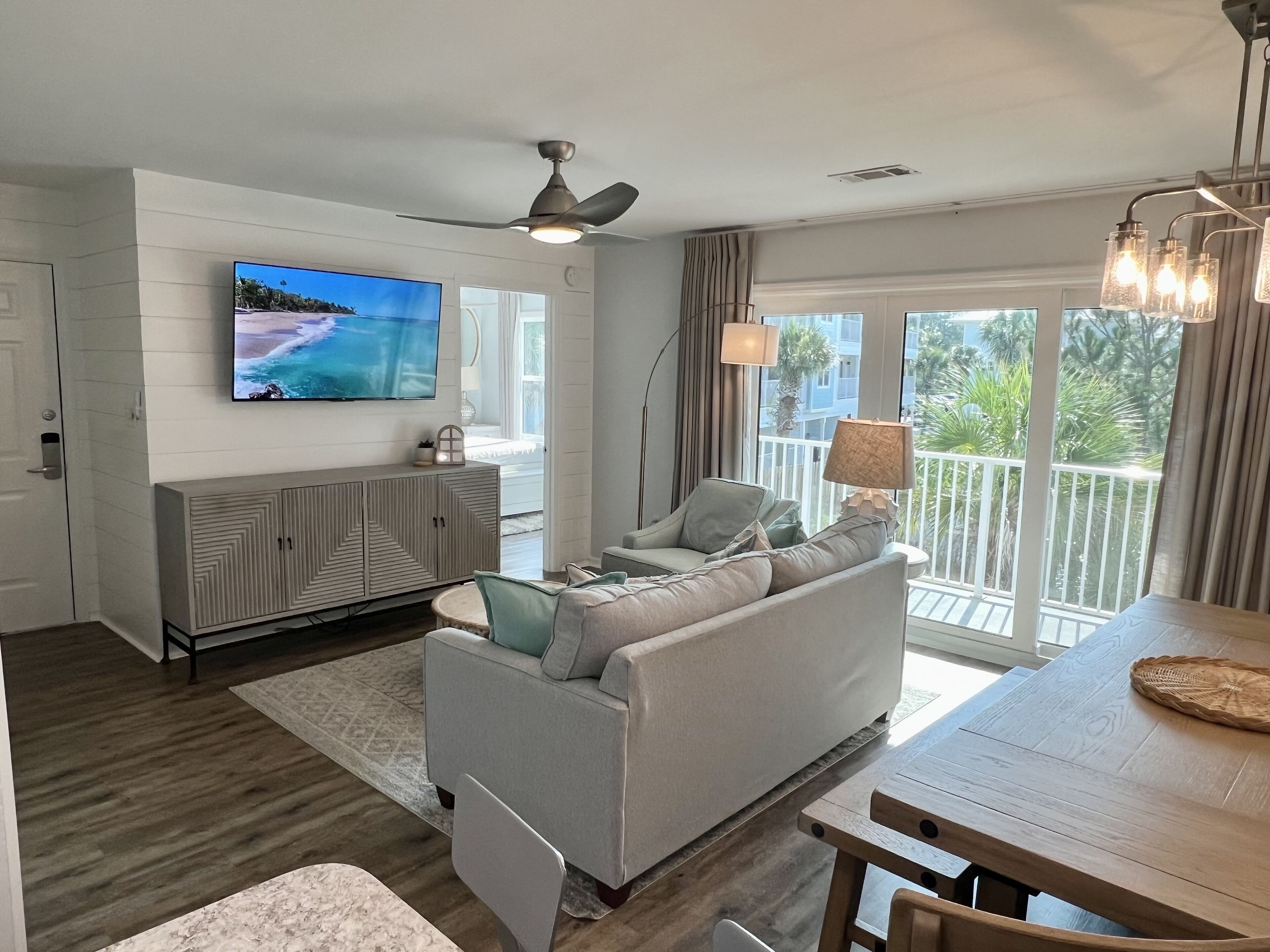 A modern living room with a flat-screen TV, a grey sofa, a wooden dining table, and a balcony with a view of greenery.