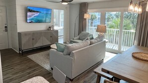 A modern living room with a flat-screen TV, a grey sofa, a wooden dining table, and a balcony with a view of greenery. - Beachside Dreams, at Boutique Beachside Villas, Newly Renovated and Decorated (Santa Rosa Beach)