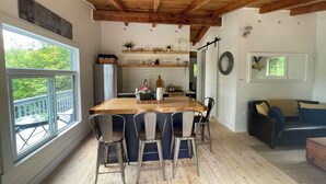 A spacious kitchen with a wooden island, a dining area with four chairs, a large window, and a view of a deck. - The Bay Stay! Fully Renovated Off-Grid Waterfront Cottage on Burridge Lake  (Godfrey)