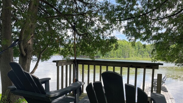 A wooden deck with Adirondack chairs overlooking a calm lake, surrounded by trees. - The Bay Stay! Fully Renovated Off-Grid Waterfront Cottage on Burridge Lake  (Godfrey)