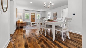 A dining area with a wooden floor, white dining table, and chairs. There is a chandelier above the table, and a door leading to another room. - A Winfield Bungalow (Dover)