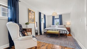 A bedroom with a bed, a chair, a bedside table, a lamp, and a painting on the wall. - A Winfield Bungalow (Dover)