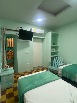Two beds with green and white bedding, a flat-screen TV, and a small nightstand with a vase of flowers.