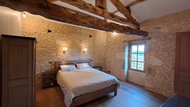 Un dormitorio con pared de piedra, vigas de madera, una cama, mesitas de noche y un ventanal.