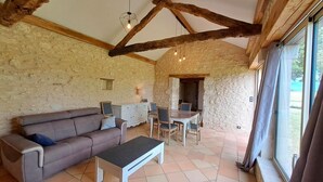 Una habitación rústica con vigas de madera a la vista, una pared de piedra, un sofá, una mesa de comedor con sillas y una ventana.