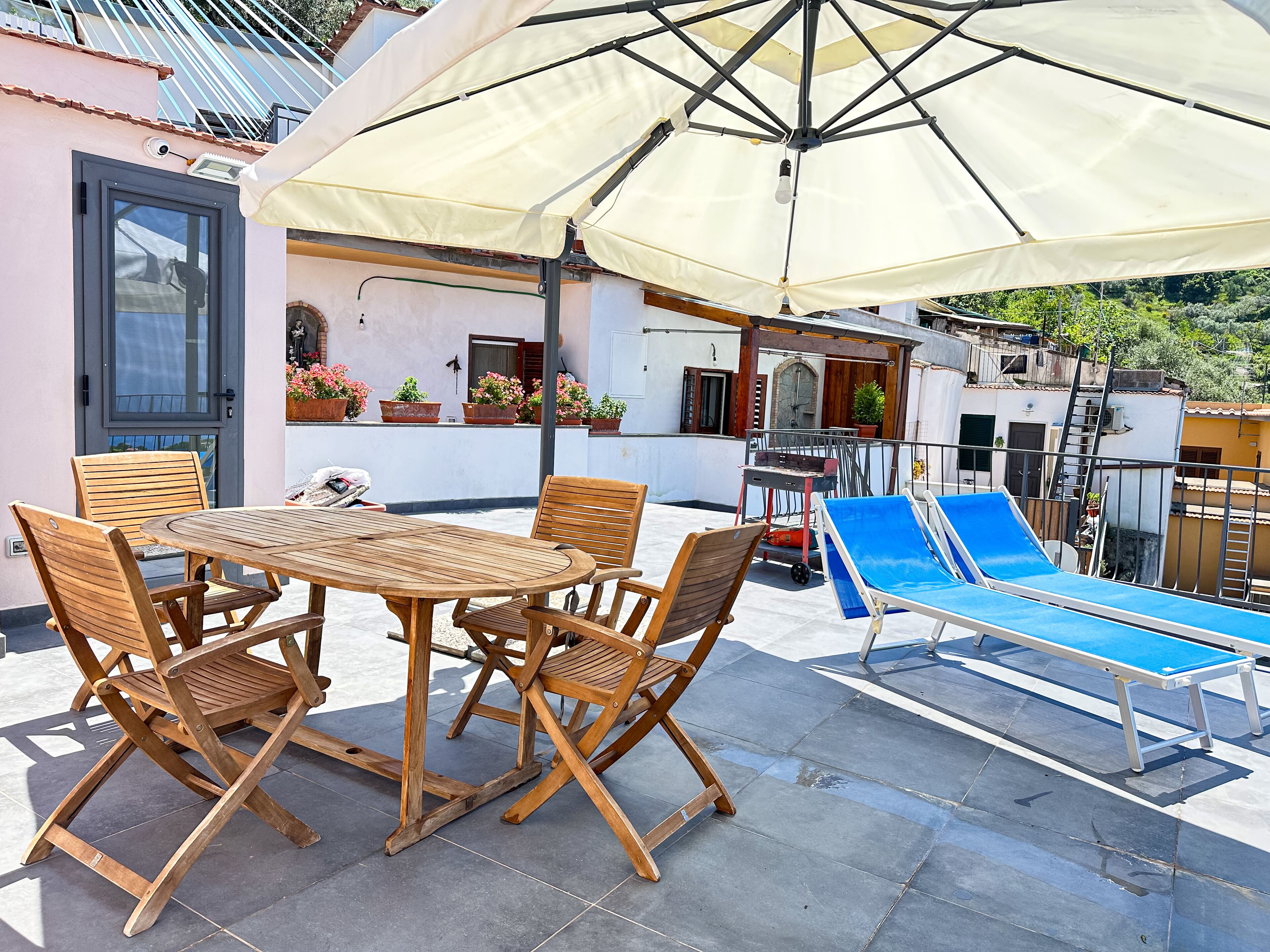 A patio with a round wooden table and four chairs, a blue sun lounger, and a large umbrella providing shade.