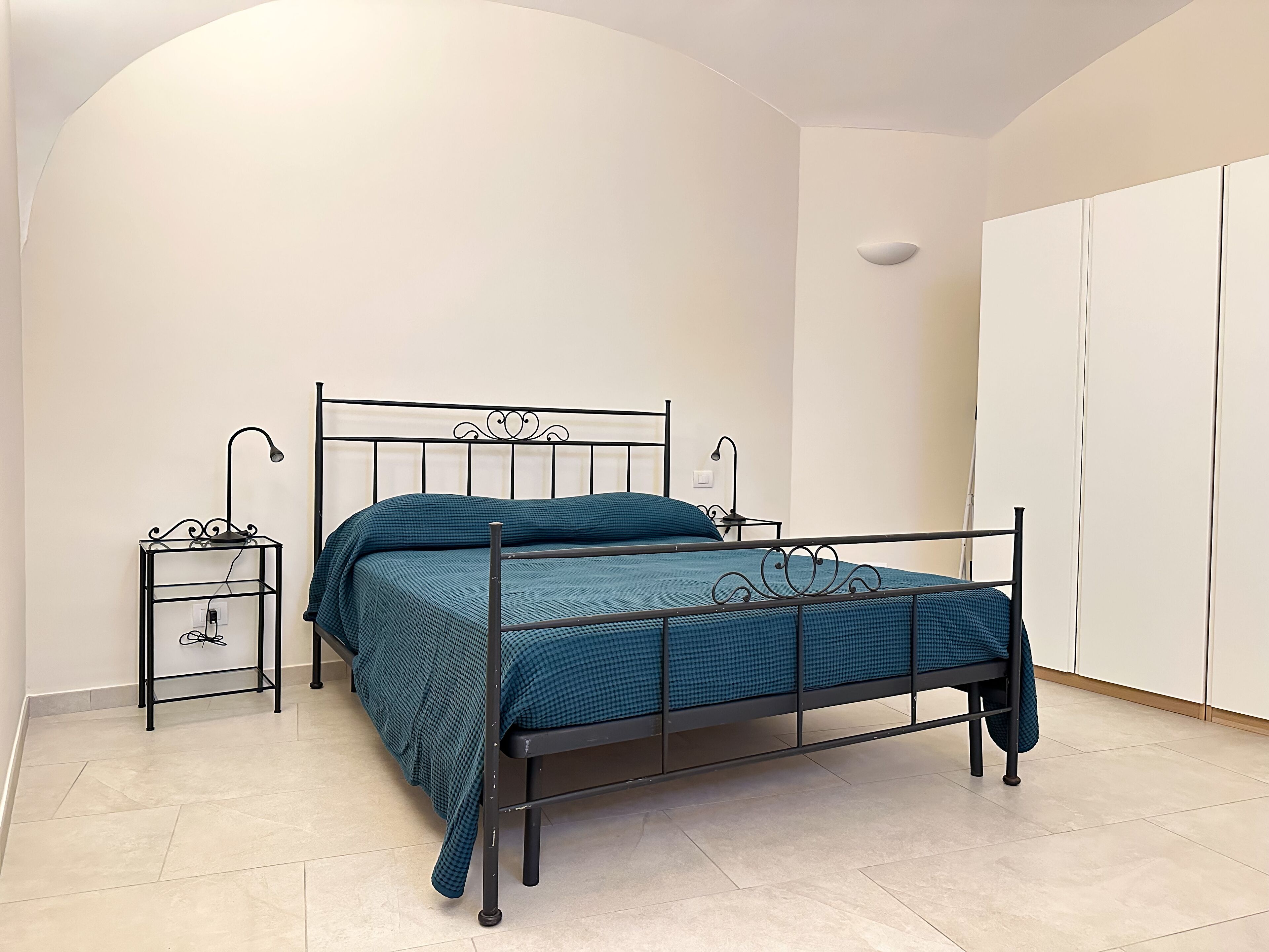 Ein Einzelbett mit Metallgestell, ein Nachttisch mit Lampe und ein Kleiderschrank in einem Zimmer mit weißen Wänden und Fliesenboden.