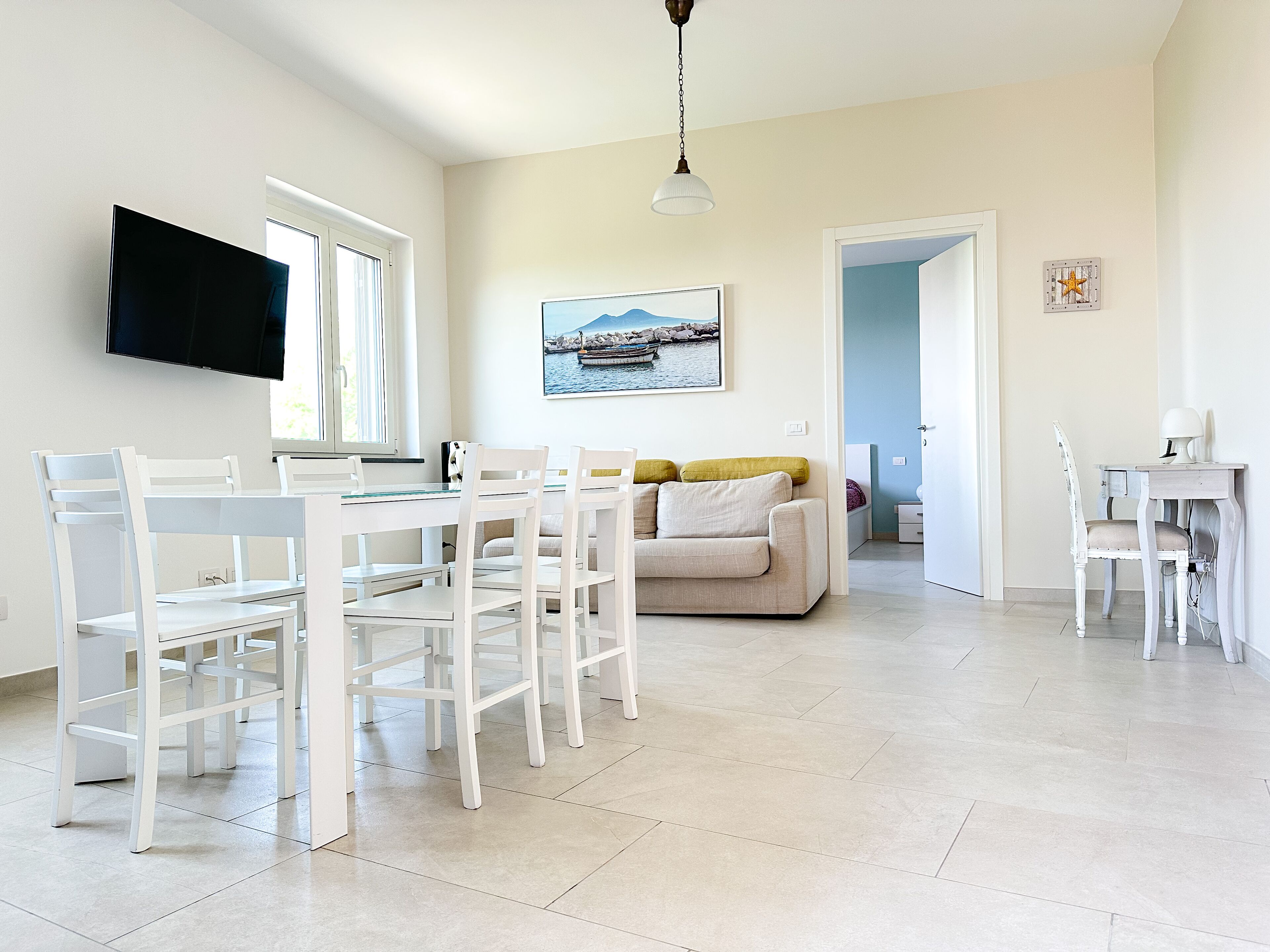Uma sala de estar clara e espaçosa, com uma mesa de jantar, cadeiras brancas, um sofá e uma televisão na parede.