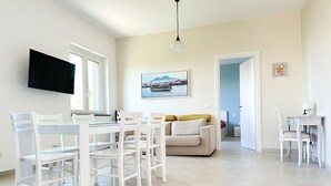 A bright, spacious living area with a dining table, white chairs, a sofa, and a television mounted on the wall. - La Campanella (Massa Lubrense)