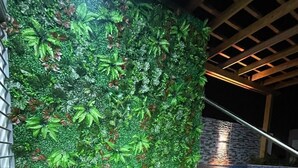Una terraza de madera con dos sillas negras y una pequeña mesa, un muro de piedra y un panel verde artificial con plantas.