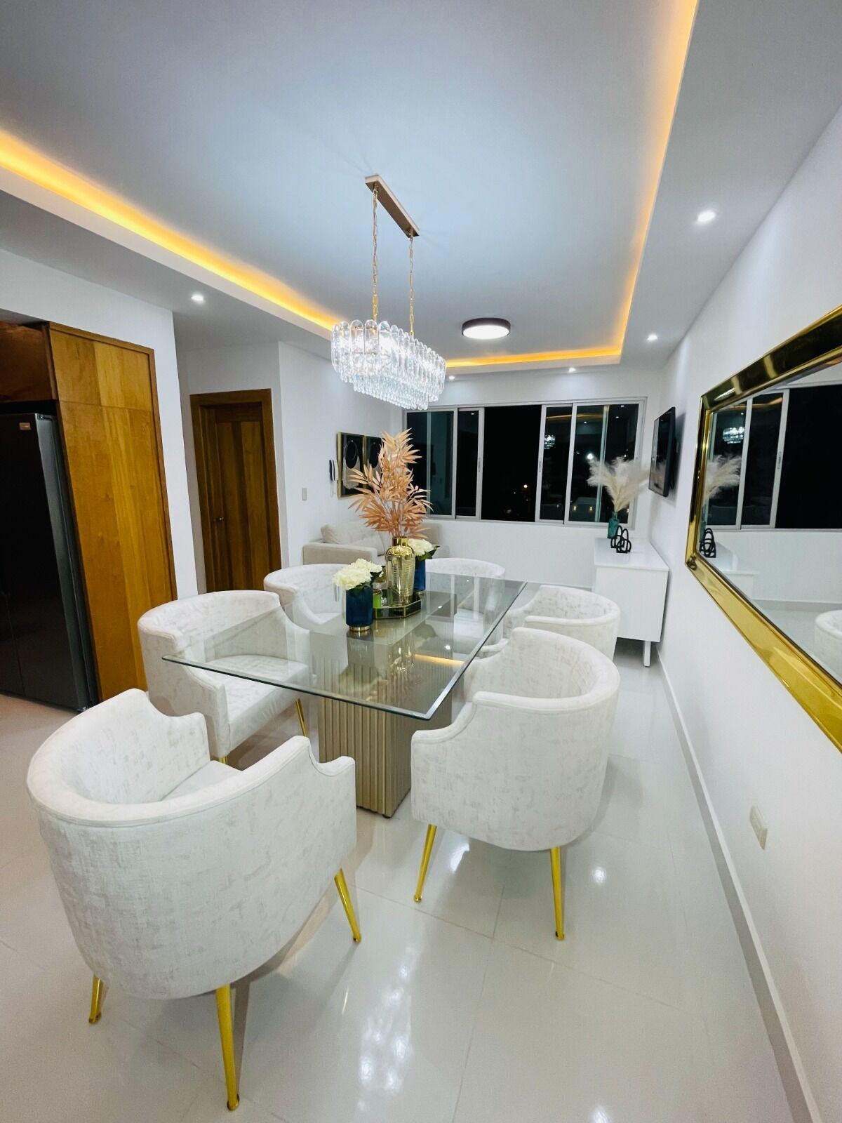 A modern dining area with a glass-top table surrounded by white chairs with gold legs, a crystal chandelier, and a large mirror on the wall.