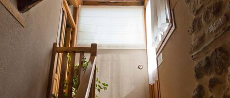 A narrow stairway with a wooden handrail, a small window with a white blind, and a stone wall on the right.