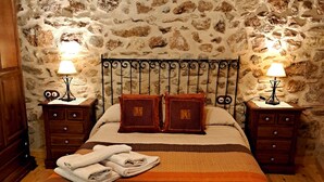 A bedroom with a stone wall, a wooden bed frame, two bedside tables with lamps, a wooden wardrobe, and a bench. - Casa La Plaza (Armuña)