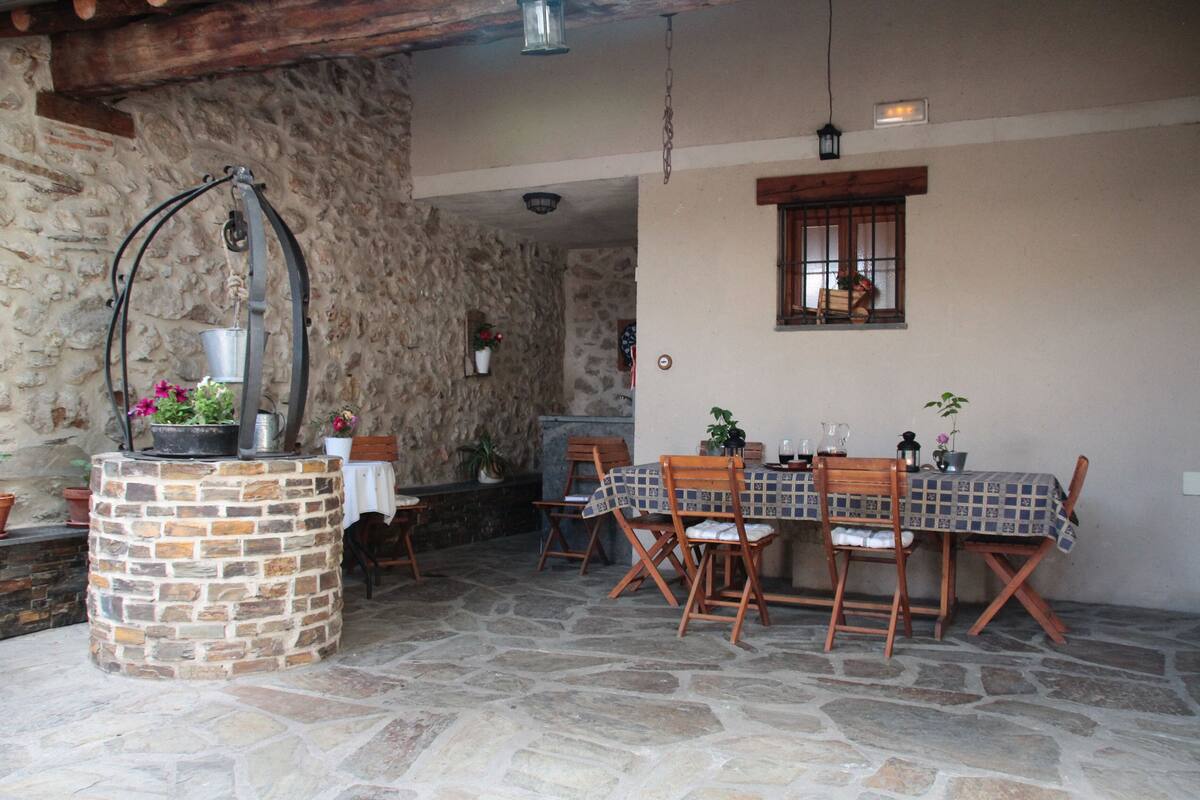 A stone-floored patio with a round table and chairs, surrounded by potted plants and a hanging metal structure.