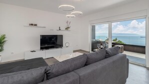 A modern living room with a large flat-screen TV, grey sofa, and a balcony with a sea view.