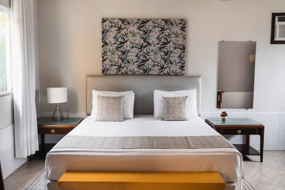 A hotel room with a bed, bedside tables, a lamp, a mirror, and a wall art piece.
