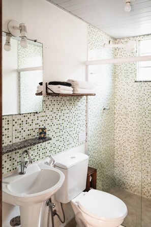 A bathroom with a sink, toilet, and shower area. There is a mirror above the sink and a towel rack with towels. The walls have a green and white patterned tile design.
