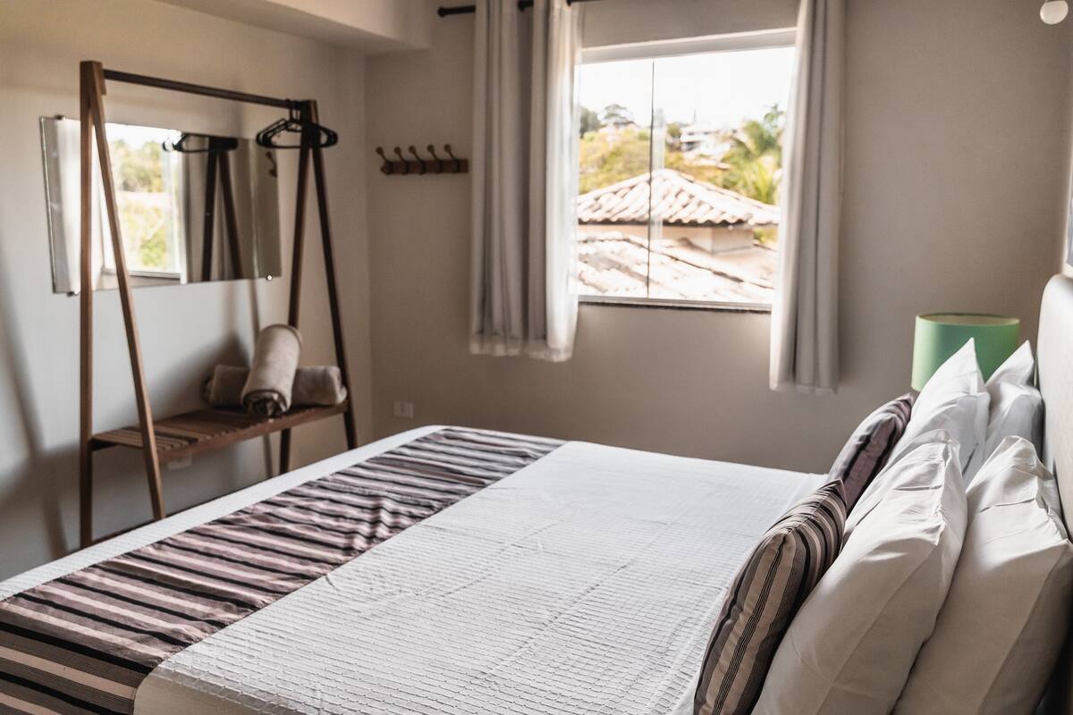 A bedroom with a bed, a wooden swing, a window with curtains, and a view of a building outside.