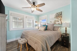 Une chambre à coucher comprenant un lit, un ventilateur de plafond, une table de chevet, une lampe et une fenêtre avec des stores.