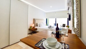 A modern hotel room with a wooden dining table, a bed with white linens, a bedside table with a lamp, and a large mirror on the wall. - OP downtown studio by Casago (Playa del Carmen)