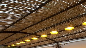 Un espacio cubierto para comer al aire libre con una mesa larga, sillas, luces colgantes y un farol verde.