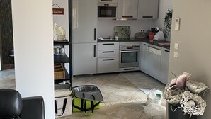 Una cocina compacta con gabinetes blancos, un microondas y una estufa. Hay un cubo de basura y una bolsa en el suelo.