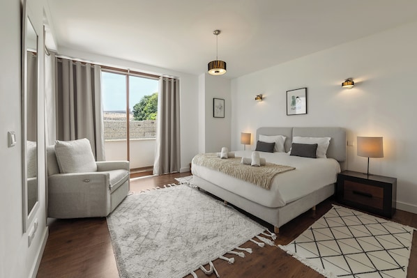 A bedroom with a bed, bedside tables, a chair, a large window, and a view of the outdoors.