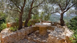 An outdoor dining area with a stone table surrounded by stone walls, set in a natural landscape with trees and hills in the background. - Cretan Stone Villa, A/C, Private Pool, Spectacular 360° Views of the Countryside, Coastline & Sea! (Stalos)