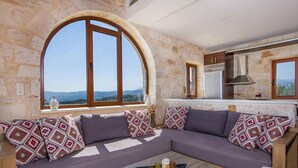 A modern living room with a stone wall, a large arched window, a sofa with patterned cushions, and a coffee table with decorative items. - Cretan Stone Villa, A/C, Private Pool, Spectacular 360° Views of the Countryside, Coastline & Sea! (Stalos)