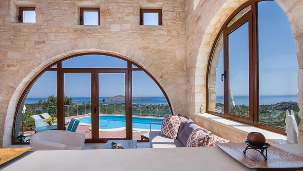 A luxurious room with a stone wall, large arched windows, a pool, and a sofa with a view. - Cretan Stone Villa, A/C, Private Pool, Spectacular 360° Views of the Countryside, Coastline & Sea! (Stalos)