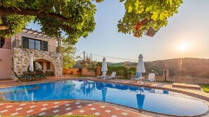 A swimming pool with a stone wall and a patio area with chairs and umbrellas. - Welcoming Family Villa - Private Pool, Stunning Mountain, Countryside & Sea Views close to Beaches! (Stalos)