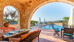 A stone archway leading to a pool area with lounge chairs and a table, overlooking a scenic landscape. - Welcoming Family Villa - Private Pool, Stunning Mountain, Countryside & Sea Views close to Beaches! (Stalos)