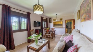 A living room with a sofa, a dining table, and a TV. There is a kitchen area with cabinets and a refrigerator. - Casa de la Cruz Iznájar by Ruralidays (Córdoba)