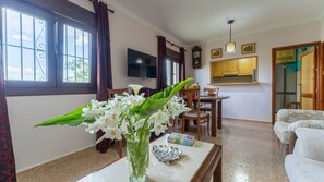Una sala de estar con comedor, un televisor montado en la pared y una cocina visible a través de una puerta abierta.