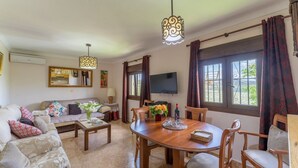 A living room with a sofa, coffee table, dining table, chairs, a TV, and large windows with curtains. - Casa de la Cruz Iznájar by Ruralidays (Córdoba)