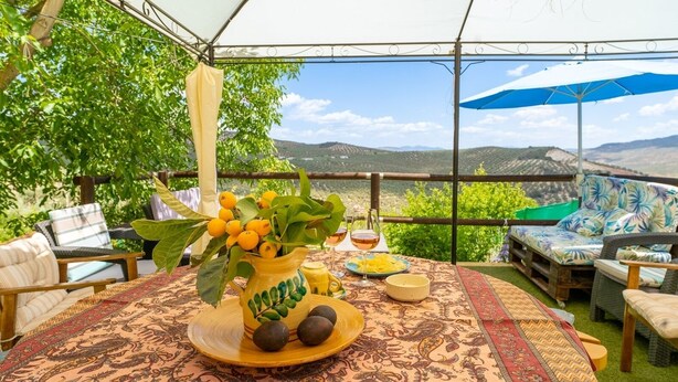 An outdoor seating area with a table set for two, featuring a pitcher of lemons and a glass of wine, surrounded by wicker furniture and a view of the countryside. - Casa de la Cruz Iznájar by Ruralidays (Córdoba)
