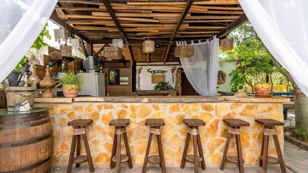 Un área de bar al aire libre de estilo rústico, con paredes de piedra, taburetes de madera y diversos objetos decorativos.