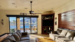 A living room with a large sofa, a coffee table, and a TV stand. There is a sliding glass door leading to a balcony with a view of the ocean and mountains.