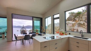 Une cuisine moderne dotée d’un grand îlot central, d’un coin repas offrant une vue sur la mer et d’une grande fenêtre donnant sur un paysage rocheux.