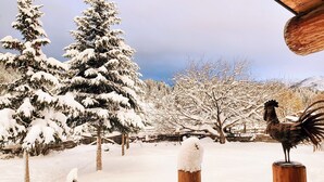 Cena externa nevada com uma estátua de galo, árvores cobertas de neve e uma construção rústica de madeira.