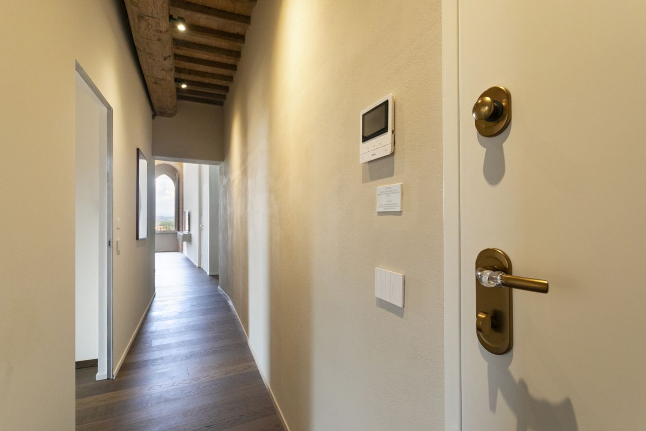 A hallway with a wooden floor, a door with a brass handle, a wall-mounted intercom, and a light switch.