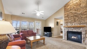 A living room with a stone fireplace, a ceiling fan, a flat-screen TV, and a large window with blinds. - Two in One! Full Condo with Lock-off Connecting Suite (Reeds Spring)