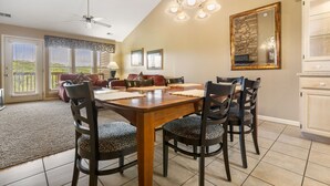 Een eetgedeelte met een houten tafel, vier stoelen en een kroonluchter. Er is een plafondventilator, een grote spiegel en een keuken met kasten.