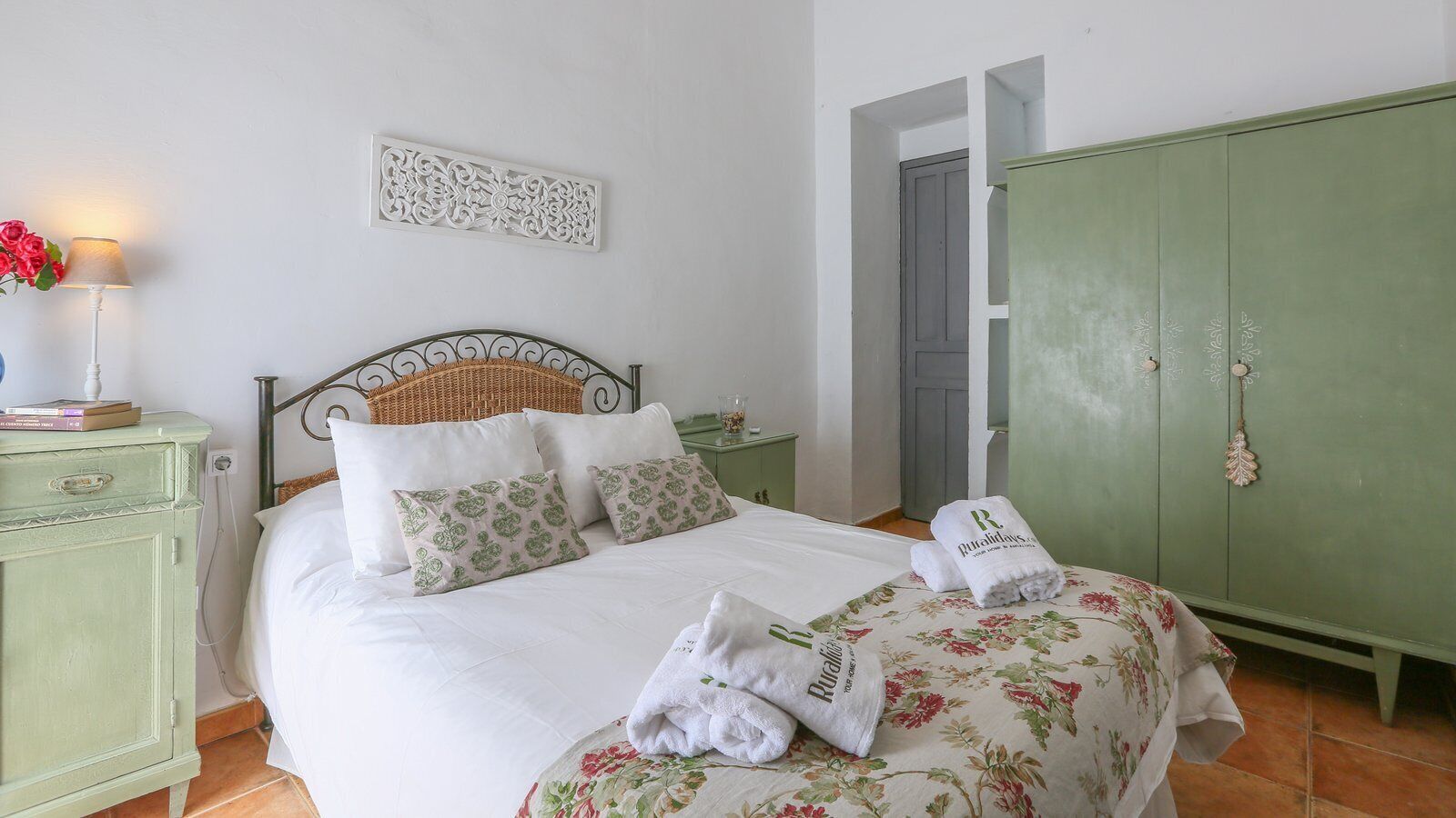 A neatly made bed with floral bedding, a green wardrobe, and a decorative wall piece.